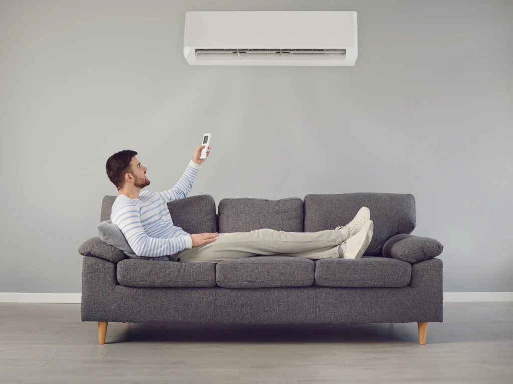 A man sitting on a couch using a remote to adjust the thermostat, enjoying the benefits of an air conditioner upgrade.